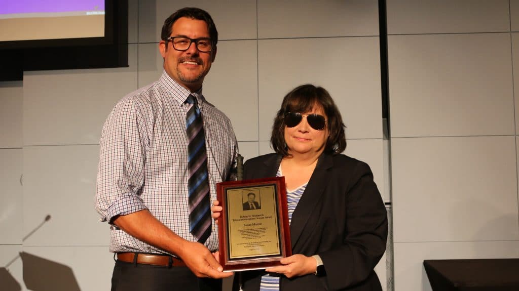 While male with dark hair and glasses wearing plaid button down shirt with tie holding a plaque with a white female with dark hair wearing sunglasses and dark jacket.