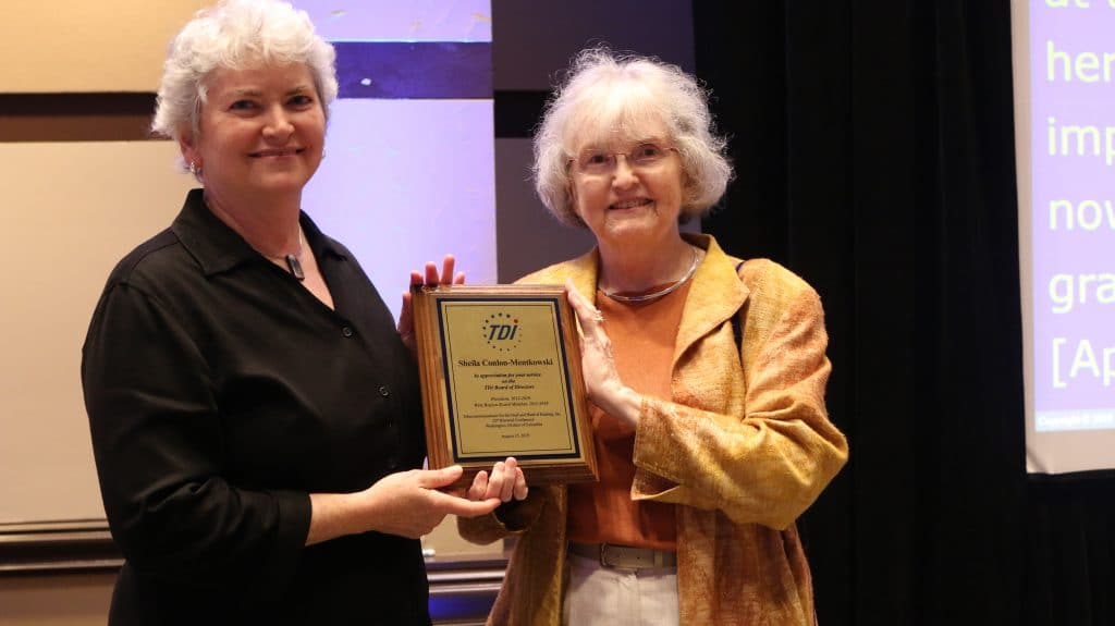 Two white women with white hair standing next to each other holding a plaque with TDI logo and text.