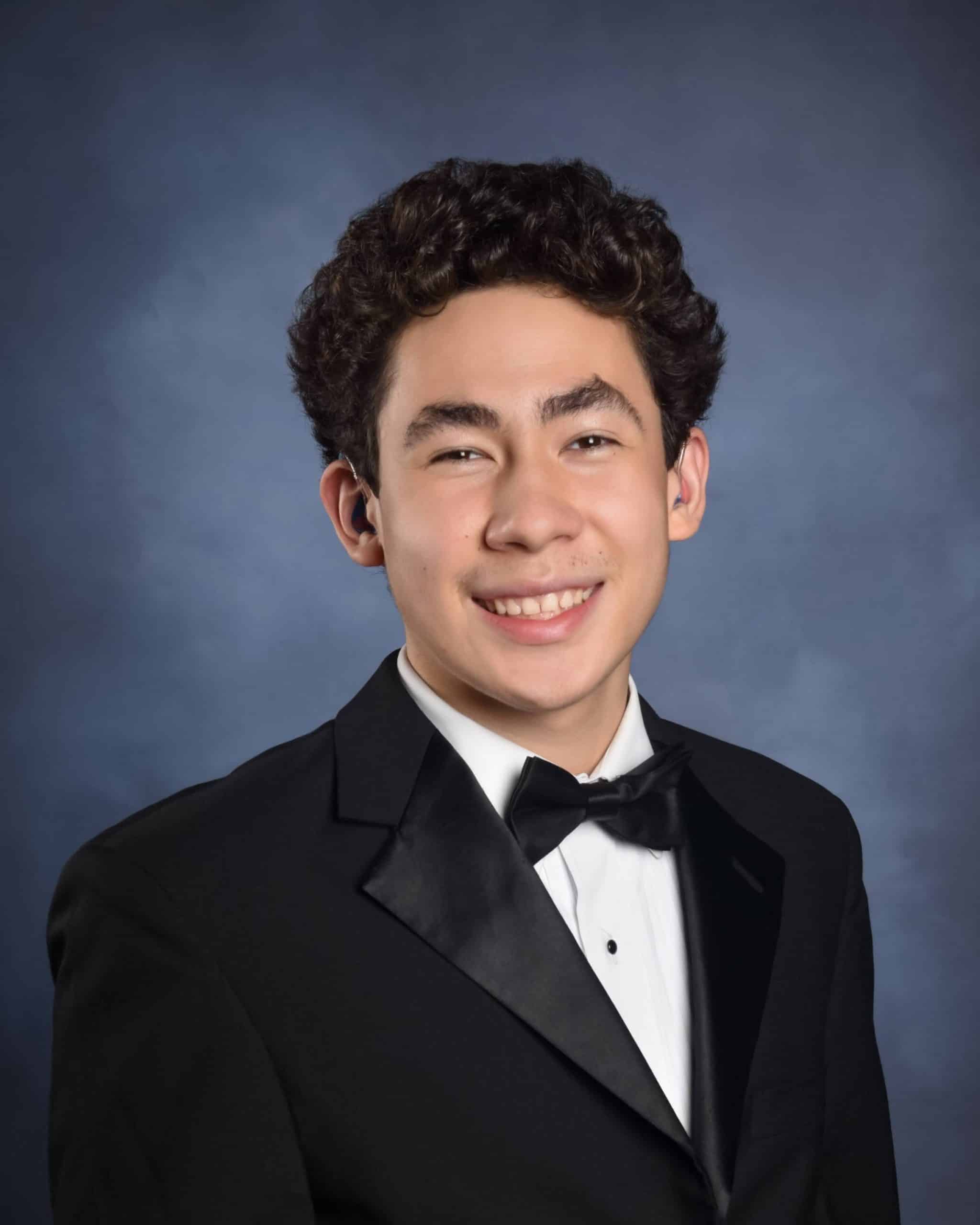 A graduation portrait of a white male in late teens. He has a thick black curly hair and wore a black tuxedo suit. He was beaming to the camera