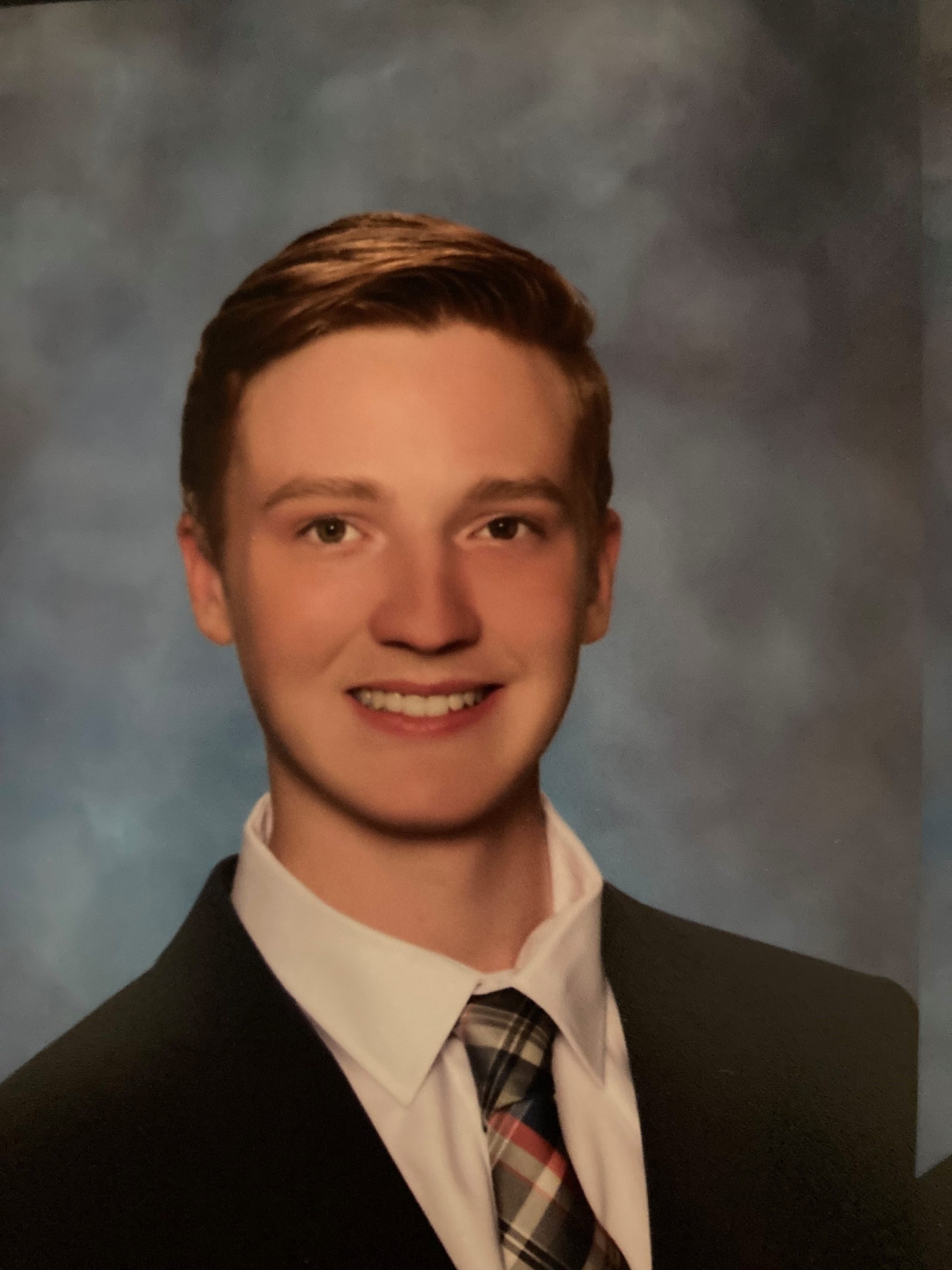 a graduation portrait of a tanned white male in late teens. He has a thick brown curly hair and wore a blue suit under the blue graduation gown with thick light blue collar.